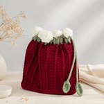 A hand-crocheted red drawstring bag styled like a flower pot, featuring white fabric flowers and green leaves at the top, with two green drawstring loops. It is displayed on a neutral cream-colored surface with dried flowers and a soft background.