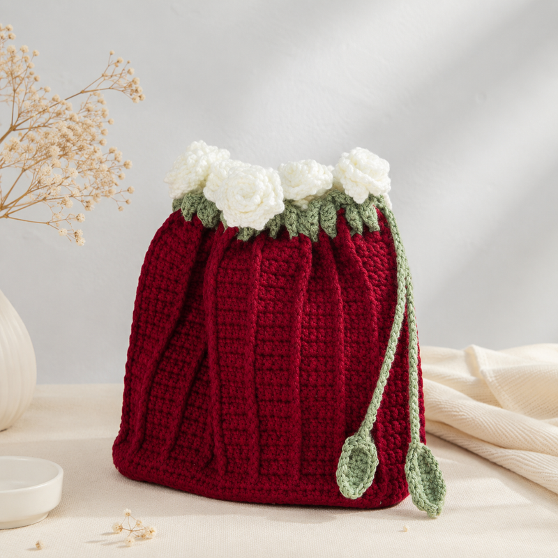 A hand-crocheted red drawstring bag styled like a flower pot, featuring white fabric flowers and green leaves at the top, with two green drawstring loops. It is displayed on a neutral cream-colored surface with dried flowers and a soft background.