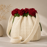 A cream-colored crocheted bucket bag with a long strap, decorated with three red crochet roses and green leaves at the top, set against a neutral beige background.